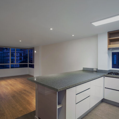 Área social de concepto abierto que integra cocina y sala, con iluminación puntual y amplios ventanales que enmarcan la vista urbana