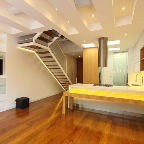 Espacio social de doble altura que integra cocina y estar, donde la escalera liviana y la iluminación arquitectónica realzan la amplitud, mientras los acabados en madera aportan calidez y sofisticación.